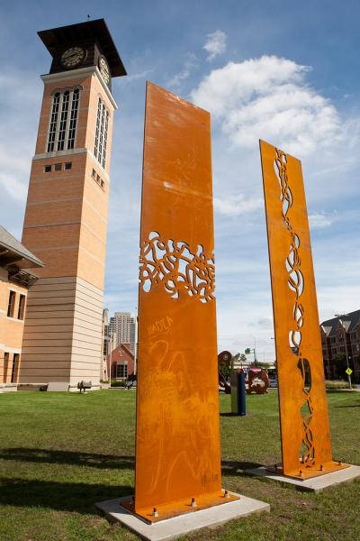 Two sculptures side by side. One has a web like design cut out through the center of the piece horizontally. The other has a section cut out vertically. The metal itself is orange in appearance.